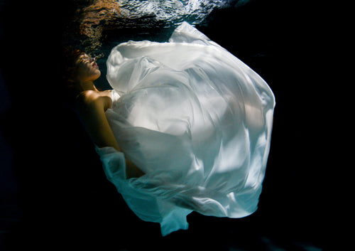 A dancer moves gracefully underwater, her body floating weightlessly as fabric flows around her. Light filters through the surface, highlighting her fluid movements and creating a sense of freedom and calm. The water becomes her stage, turning each gesture into a moment of quiet beauty.