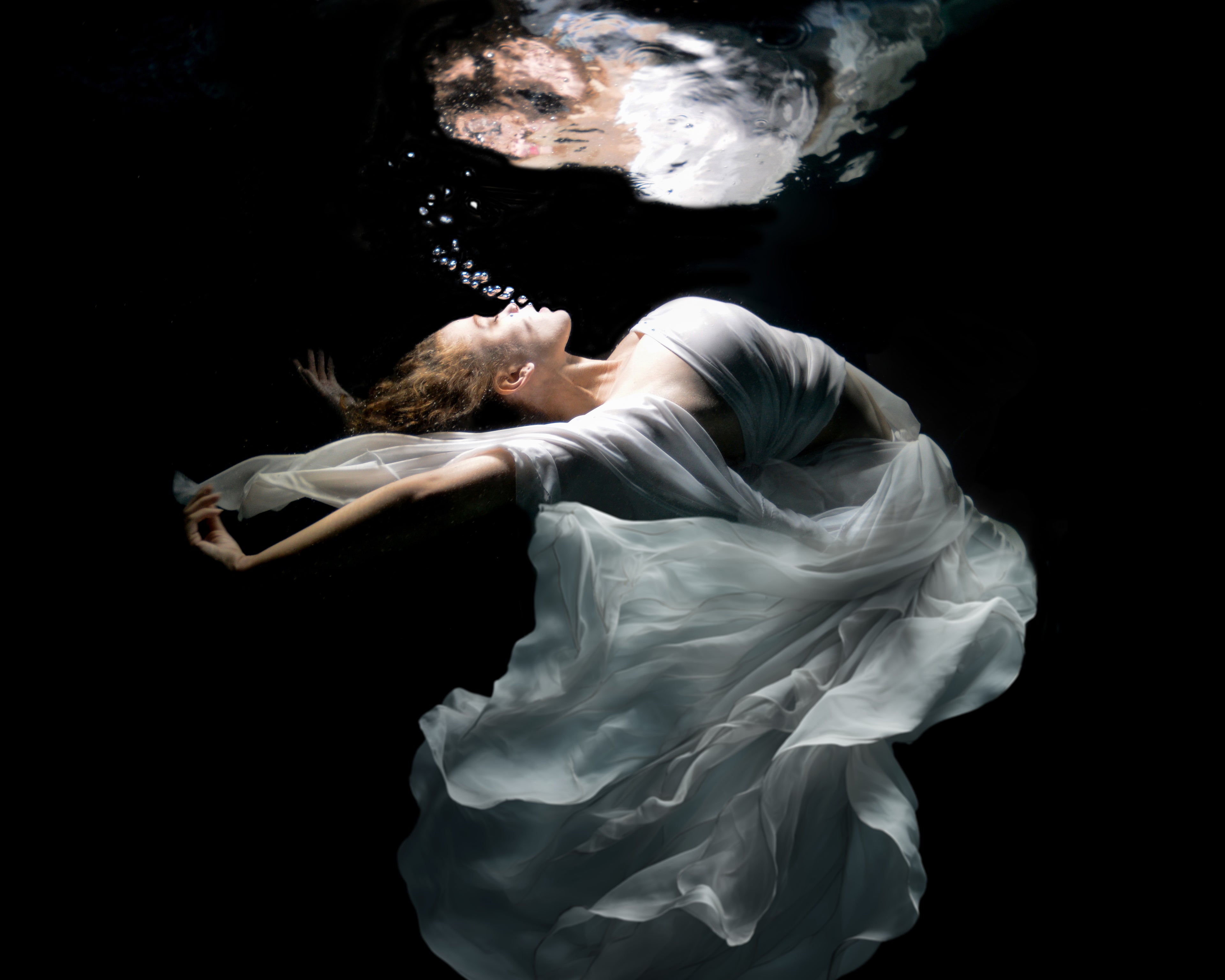 A dancer moves gracefully underwater, her body floating weightlessly as fabric flows around her. Light filters through the surface, highlighting her fluid movements and creating a sense of freedom and calm. The water becomes her stage, turning each gesture into a moment of quiet beauty.