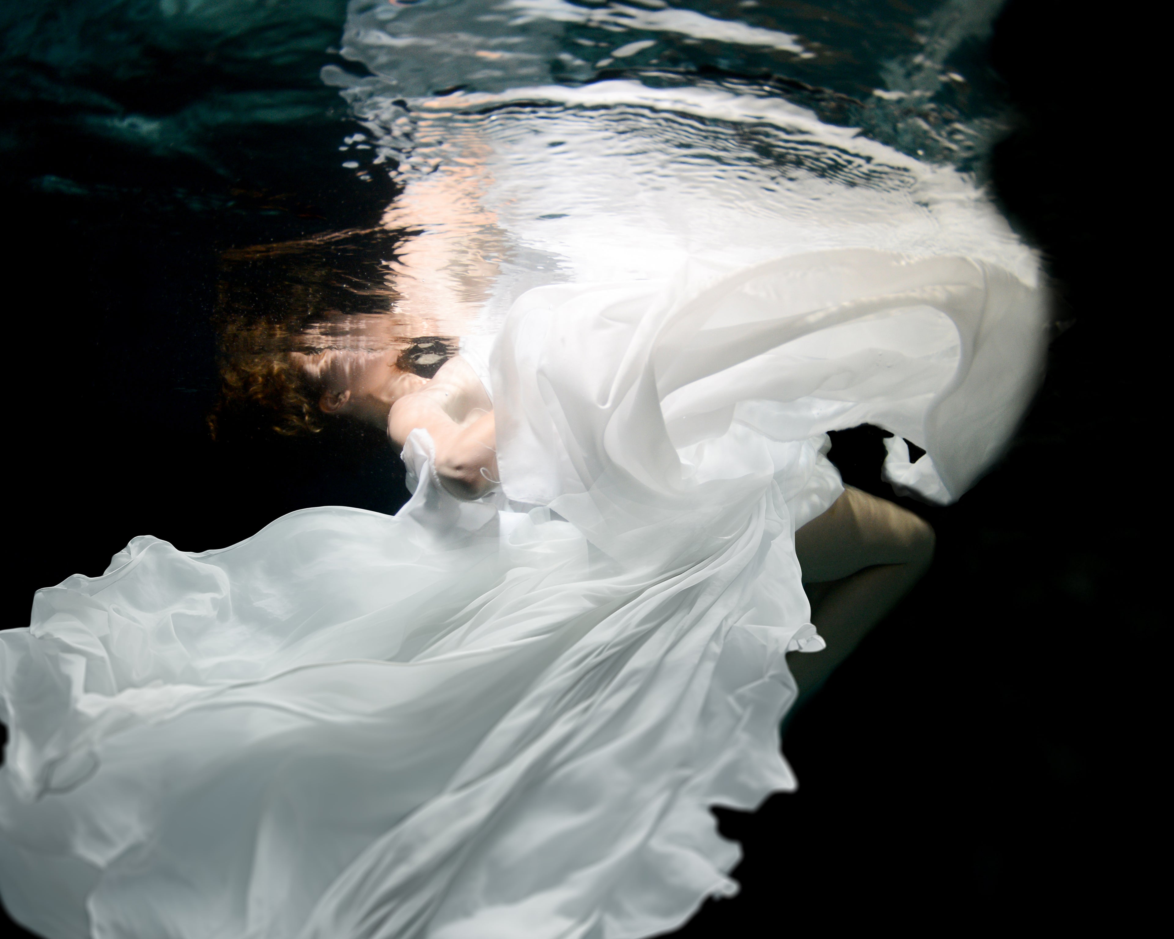 A dancer moves gracefully underwater, her body floating weightlessly as fabric flows around her. Light filters through the surface, highlighting her fluid movements and creating a sense of freedom and calm. The water becomes her stage, turning each gesture into a moment of quiet beauty.